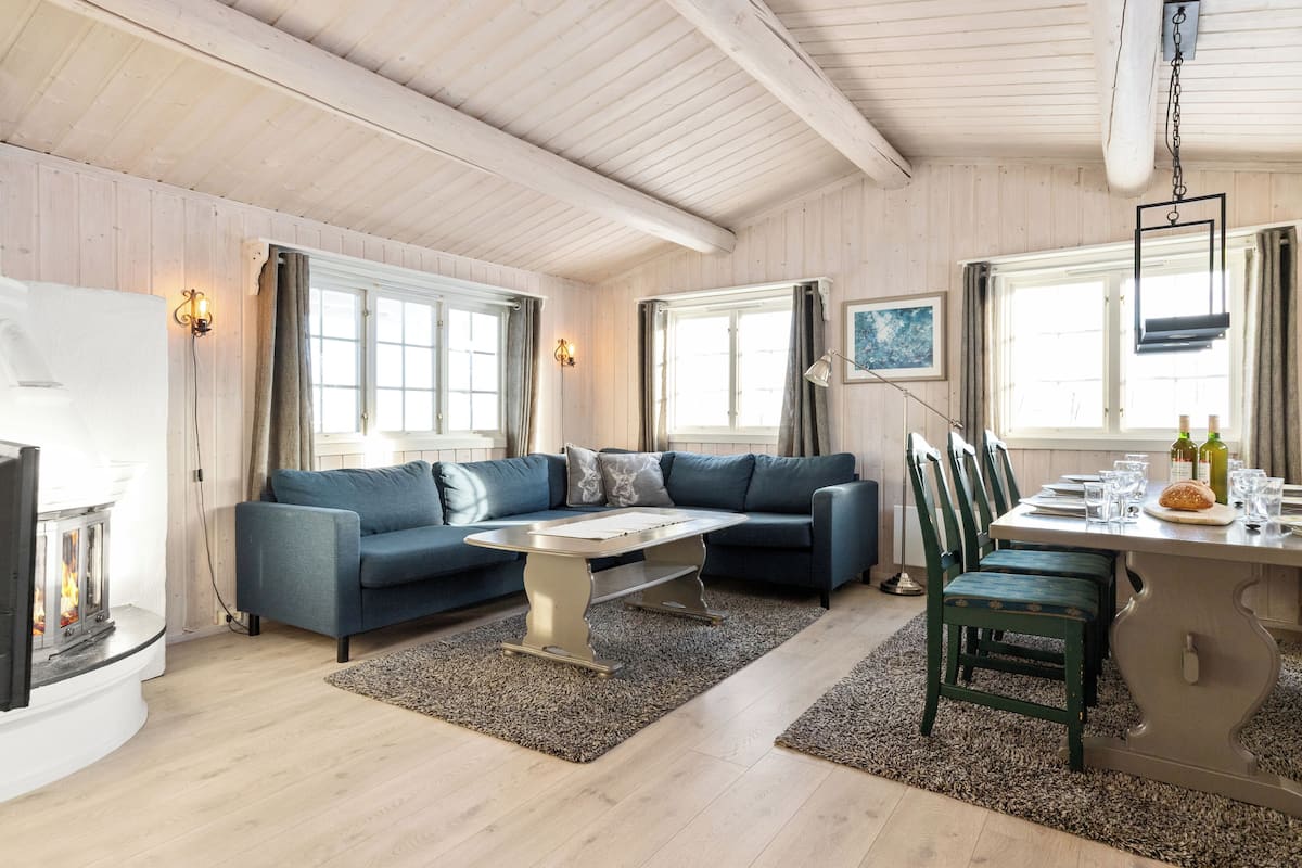 Een knus vertrek met een open haard, een blauwe bank, een eettafel met stoelen en een groot raam.