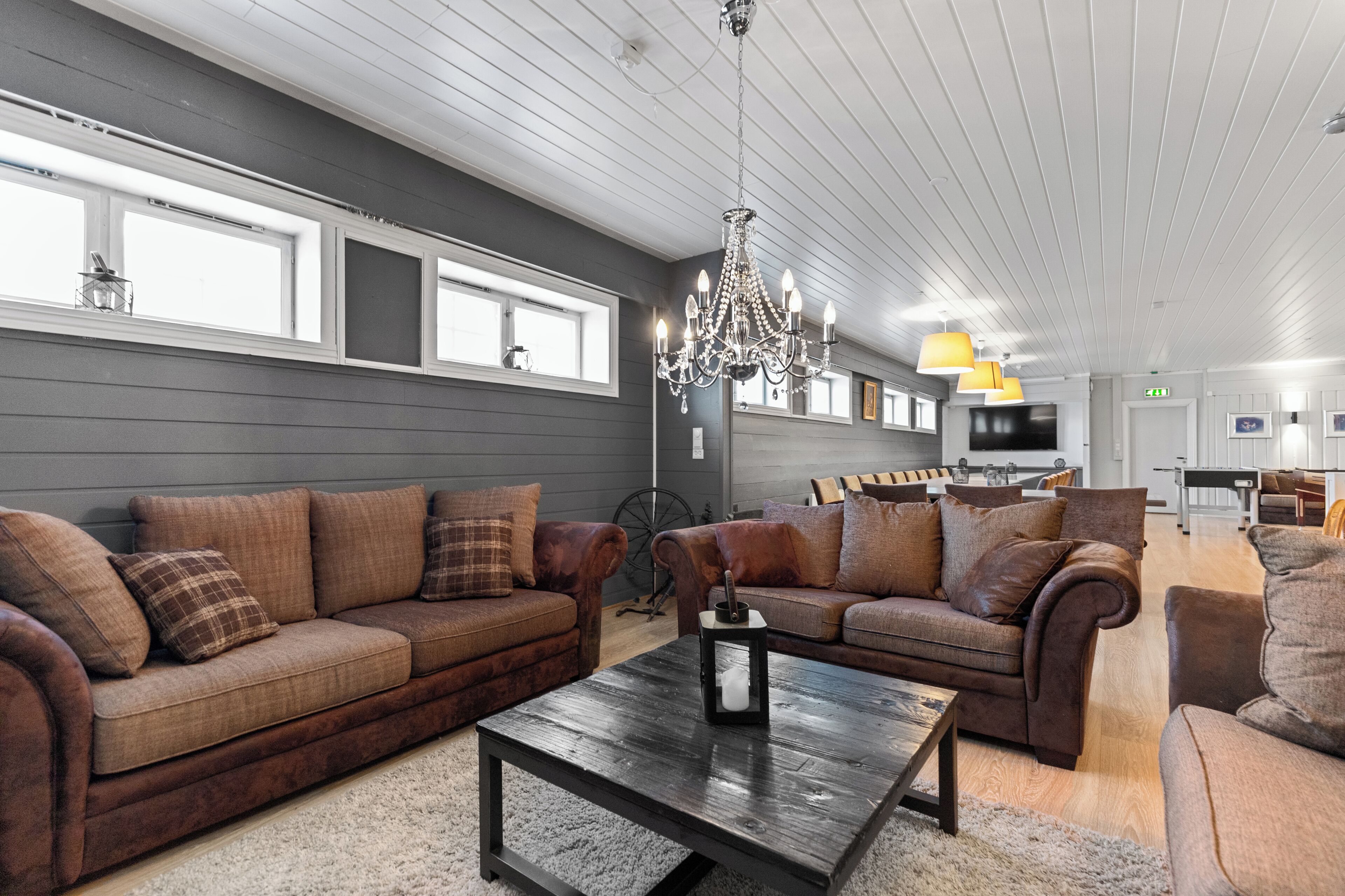 A modern hotel room with two brown leather sofas, a wooden coffee table, a chandelier, and a kitchen area in the background.
