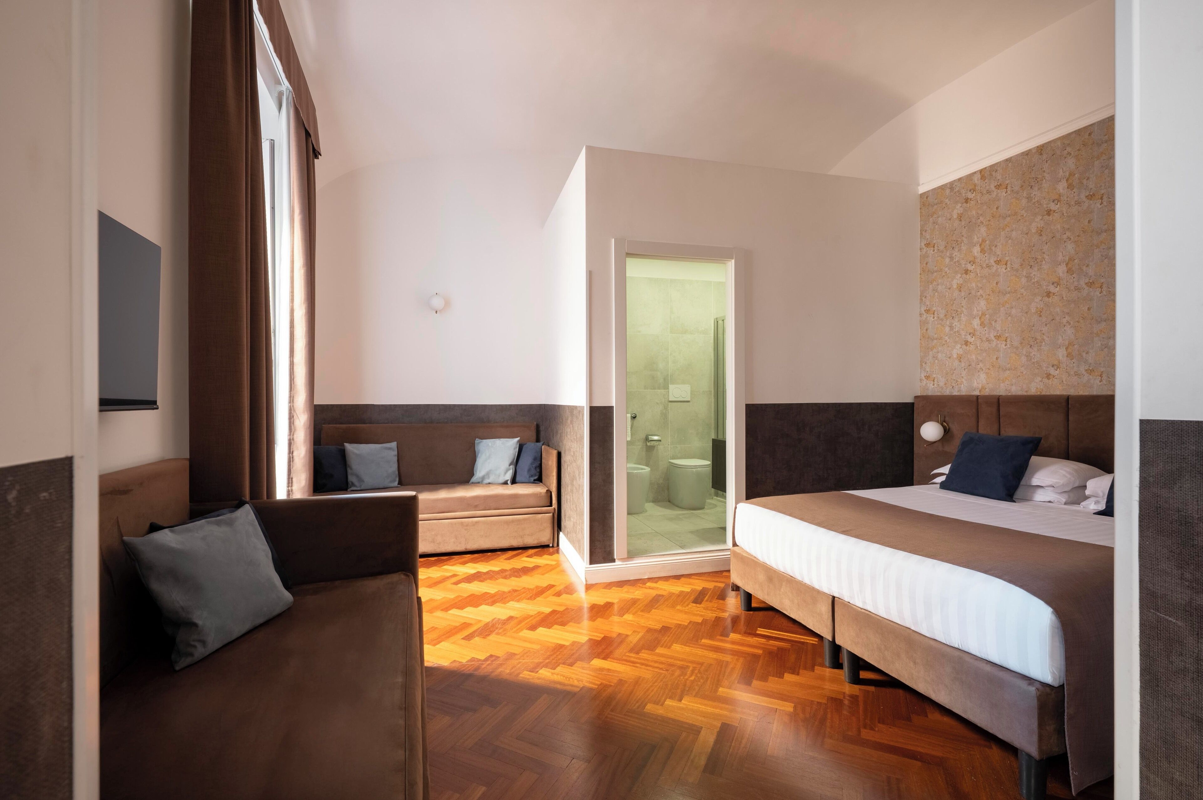 A hotel room with a wooden floor, a sofa, a bed, and a bathroom with a glass door.