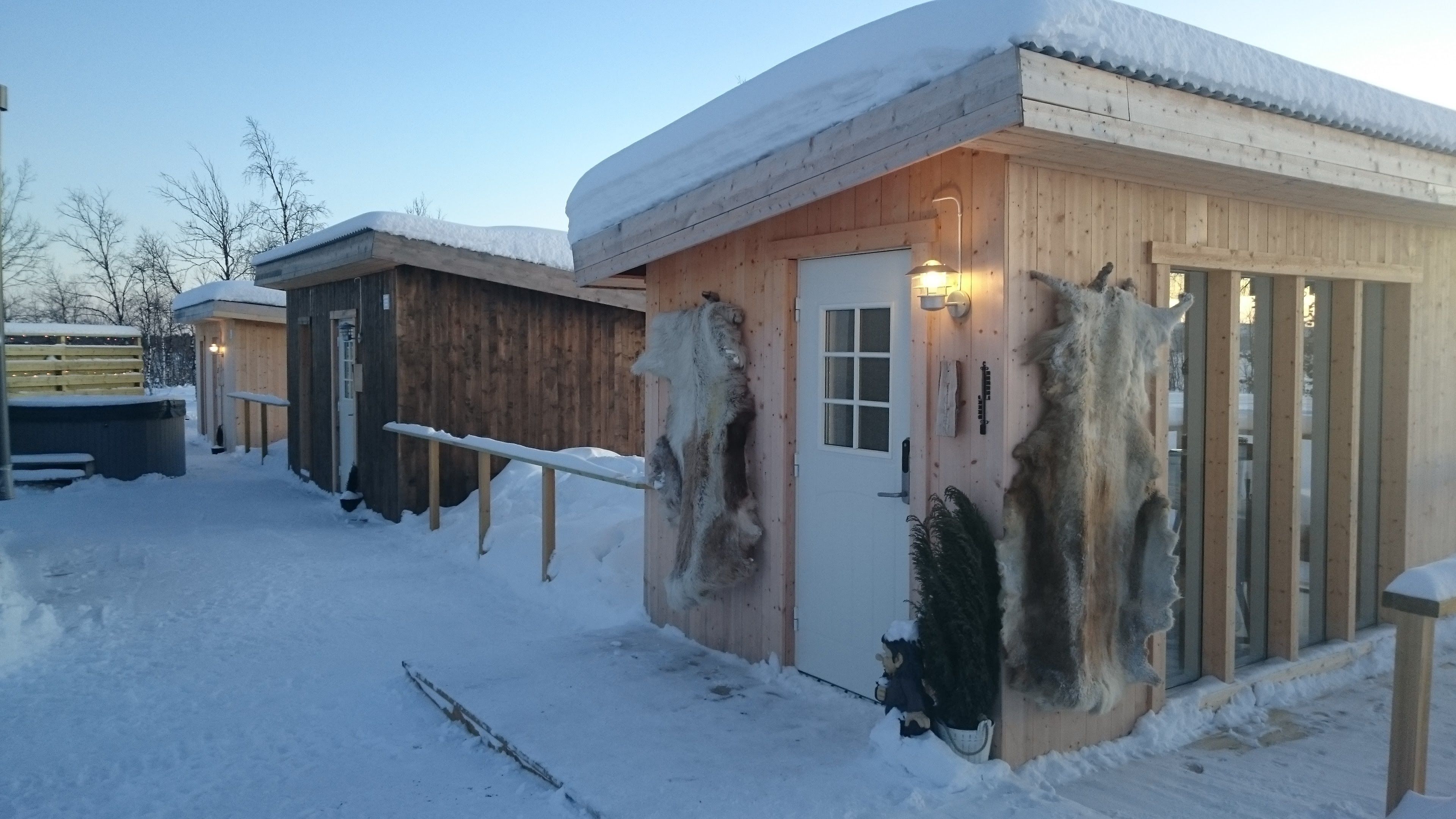 Una cabaña de madera con techo cubierto de nieve, una puerta con ventana y un accesorio de luz en la pared.