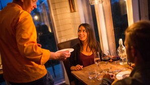 A group of people seated at a wooden table with wine glasses and a bottle, in a dimly lit room with a window. - Arctic gourmet cabin (Kiruna)