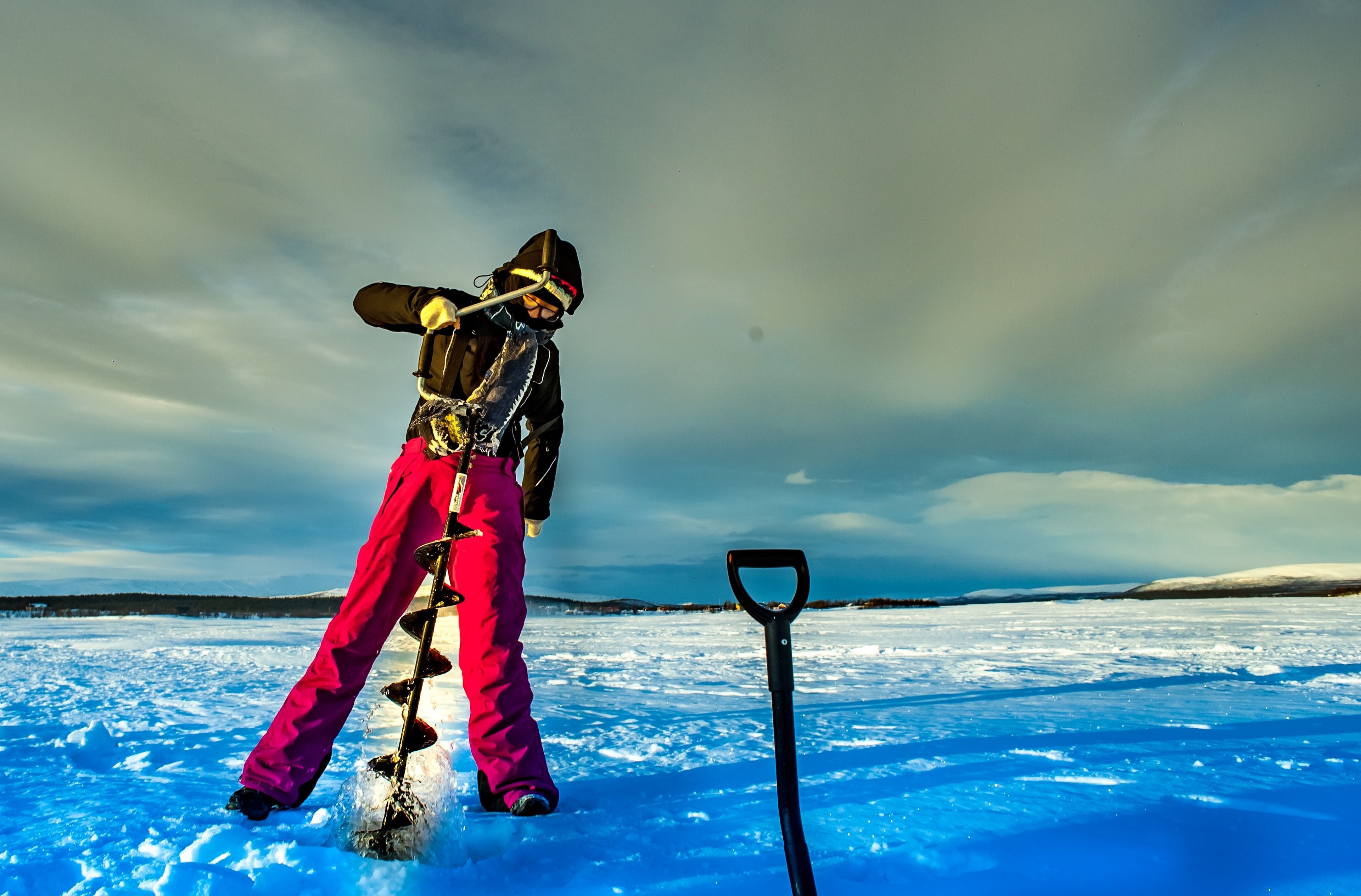 A person in pink ski gear shoveling snow on a snowy field with a shovel.