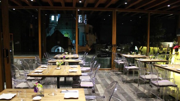 Un restaurante con mesas y sillas de vidrio transparente, techo de madera y vista a un edificio al fondo.