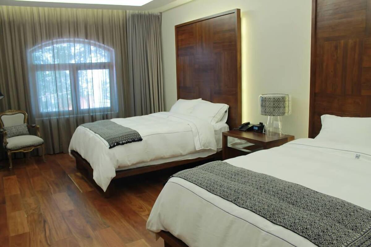 A hotel room with two beds, a wooden headboard, a chair, a lamp, and a window with curtains.