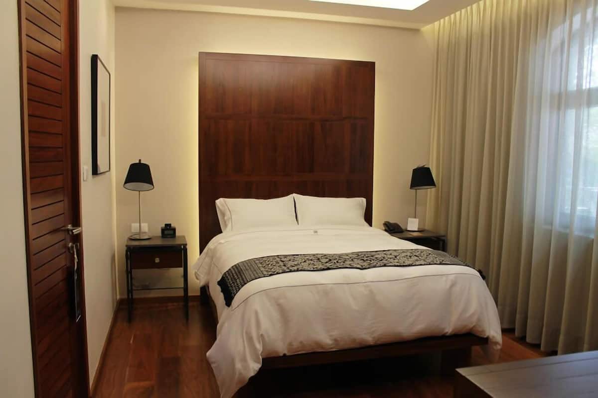 A hotel room with a wooden headboard, a bed with white linens, two bedside tables with lamps, and a window with curtains.