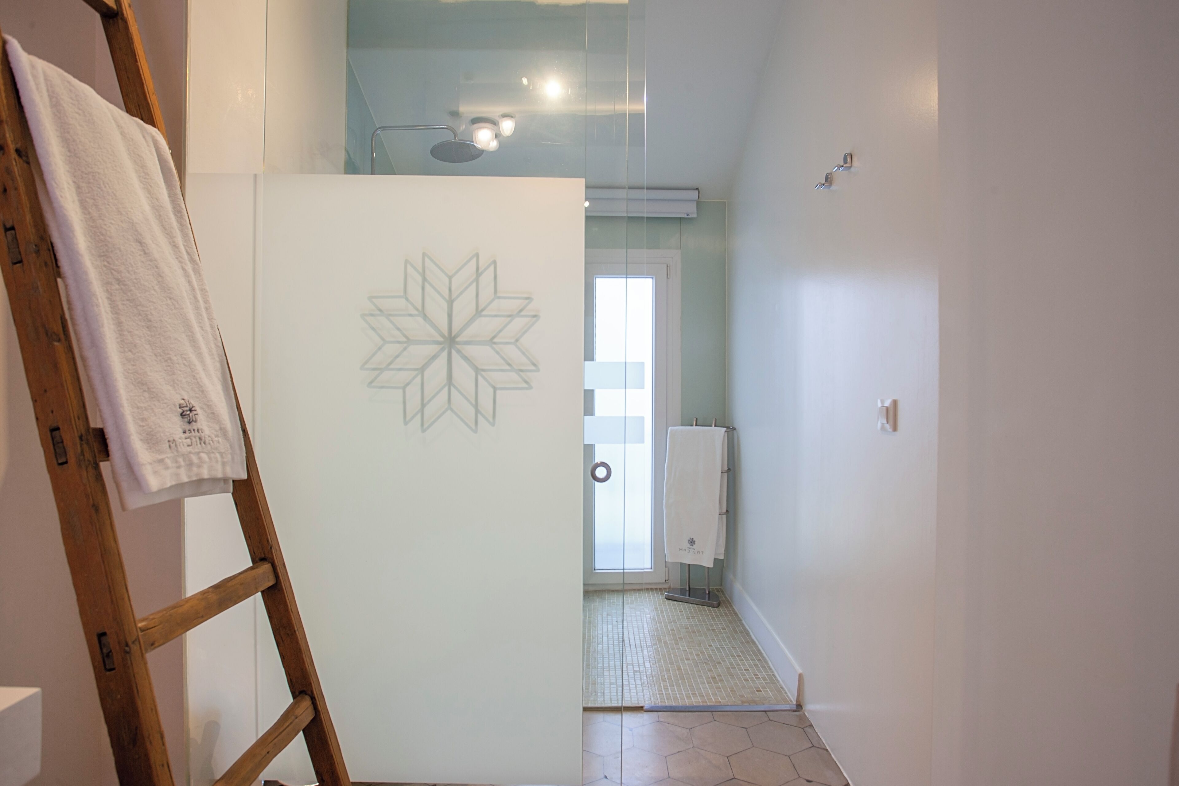 Um corredor com uma escada de madeira, uma porta branca com design geométrico, uma toalha pendurada na escada e um banheiro com piso ladrilhado e porta-toalhas.