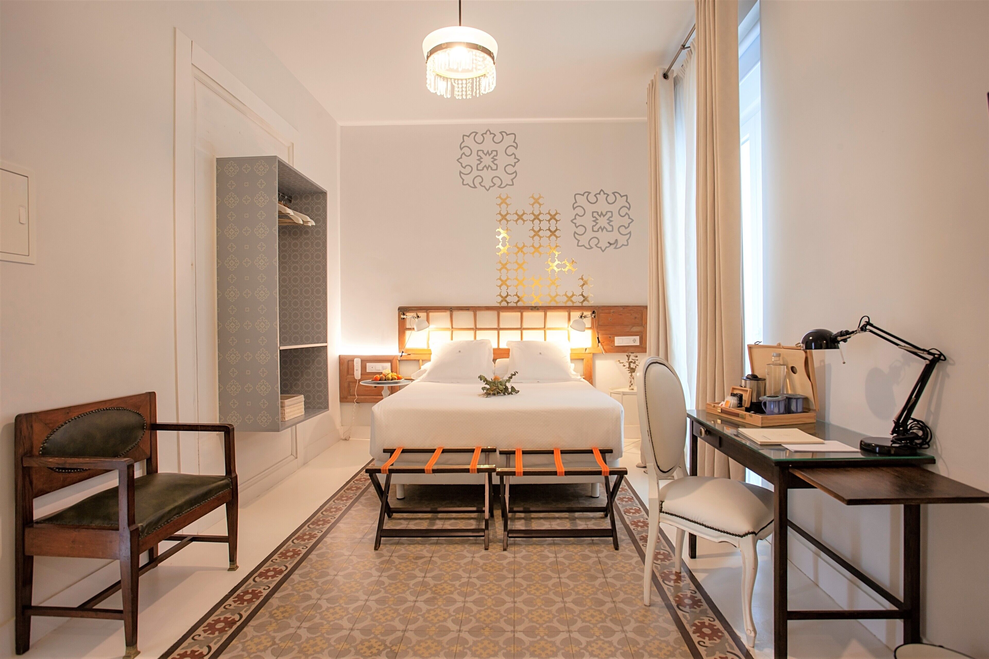 Une chambre d’hôtel comprenant un lit, un bureau, une chaise, un banc, une armoire et un lustre.