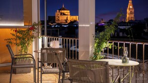 A balcony with wicker chairs and a table, overlooking a cityscape with illuminated buildings.