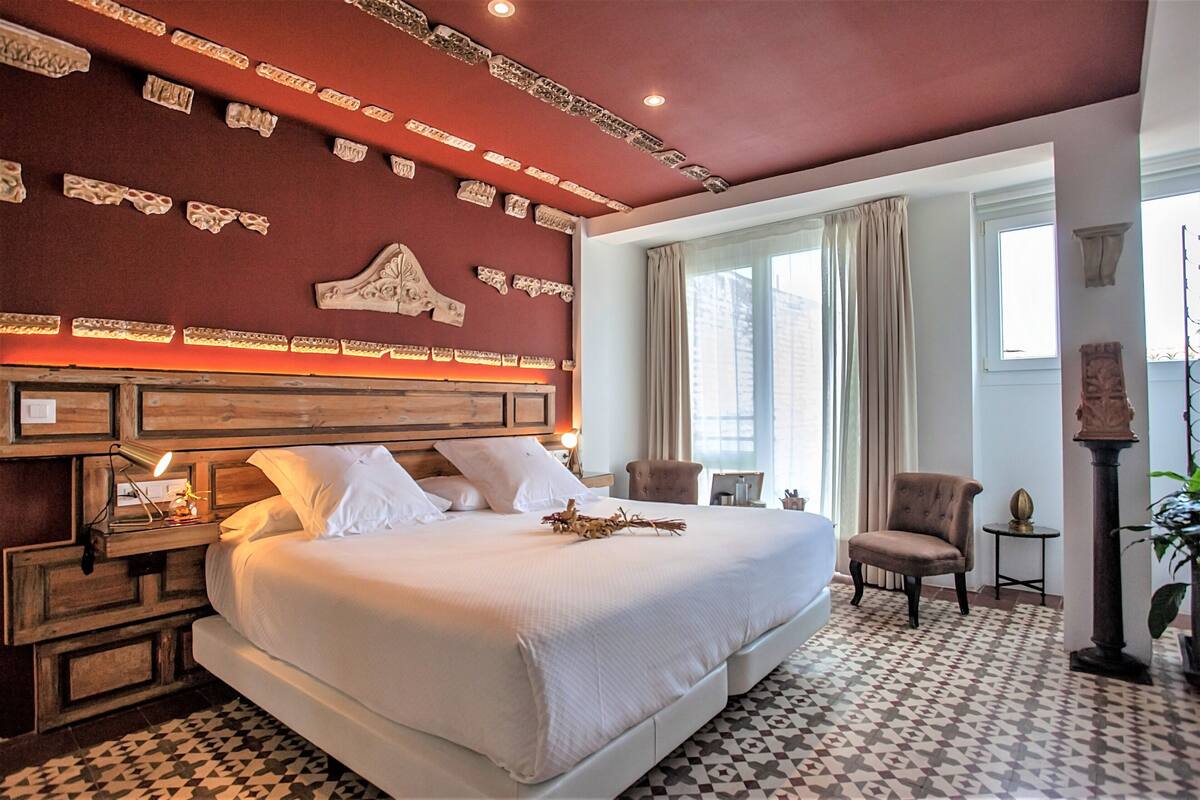 A hotel room with a large bed, wooden headboard, decorative wall panels, a small table with a lamp, a chair, and a potted plant.