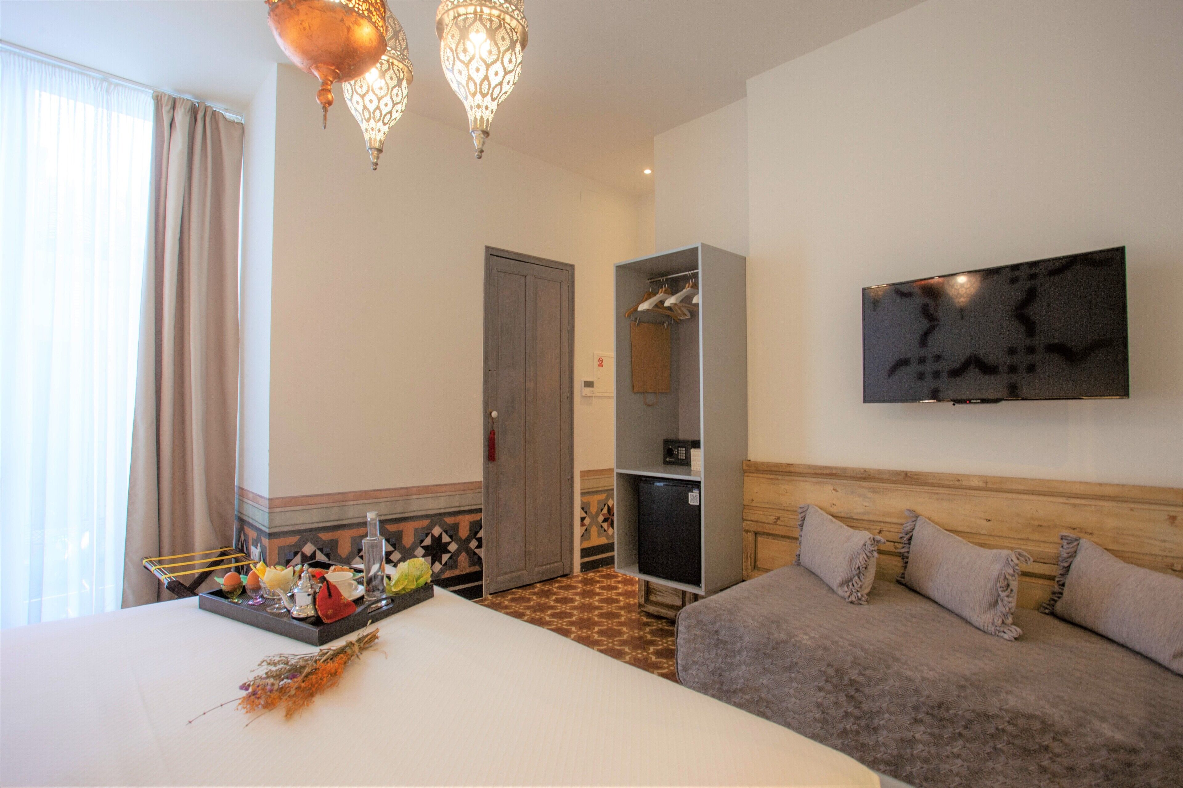 A hotel room with a bed, a sofa, a TV mounted on the wall, a wardrobe, and a decorative chandelier.