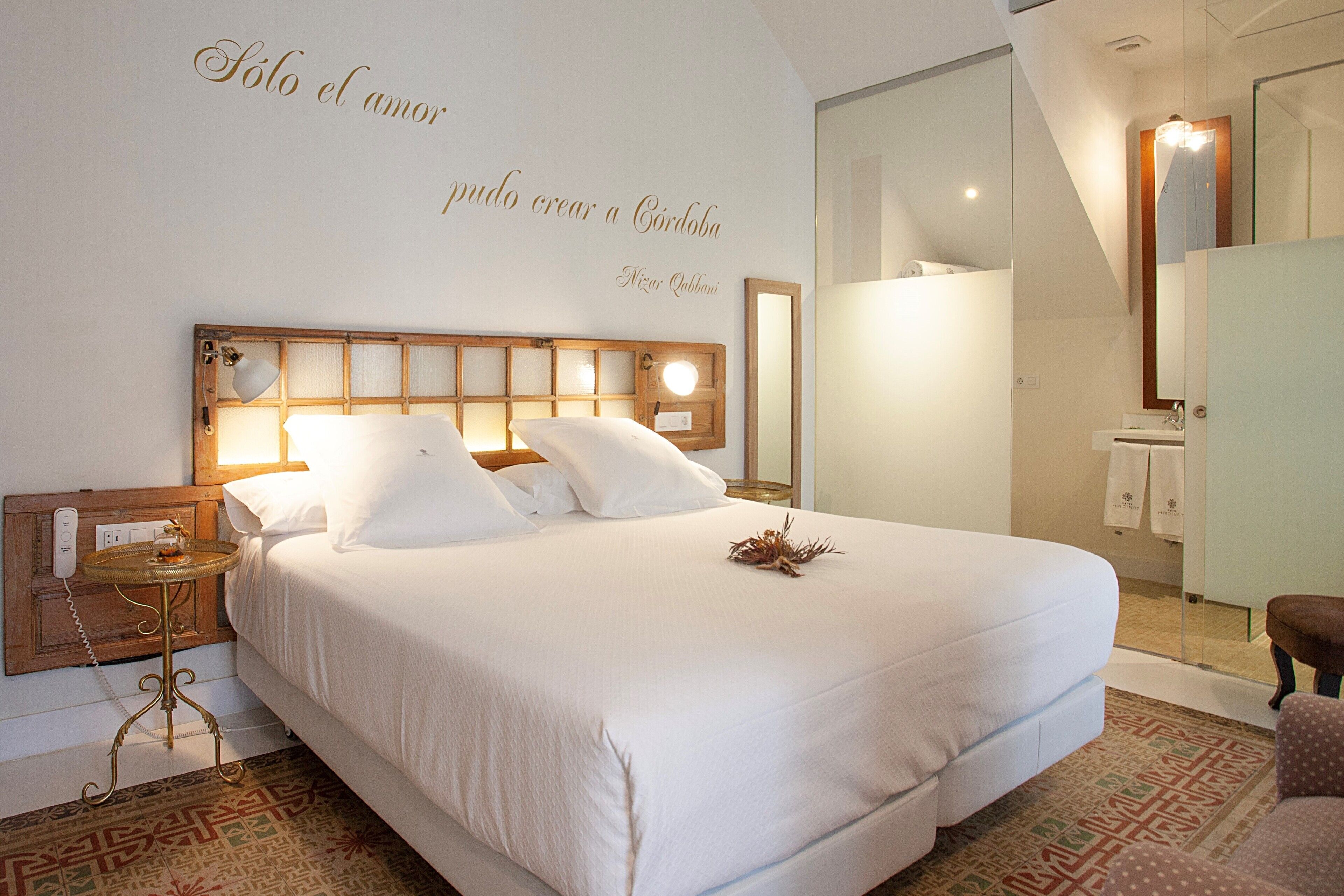 Una cama bien hecha con sábanas blancas, una mesita de noche con una lámpara y una pared con una cita decorativa.
