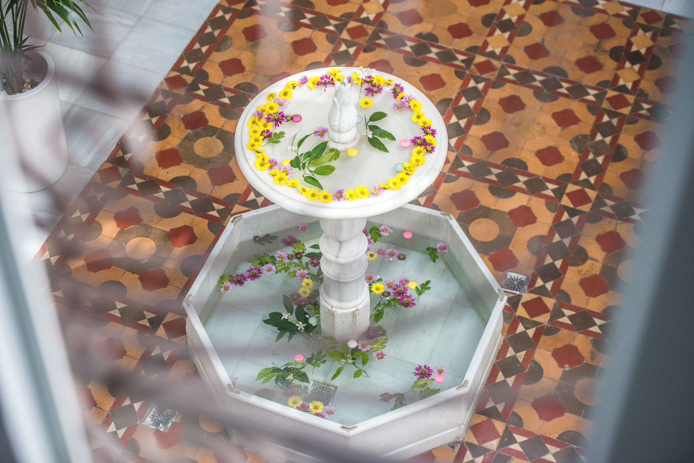 A fountain with a white pedestal and a circular basin, surrounded by yellow and purple flowers, set on a patterned floor.
