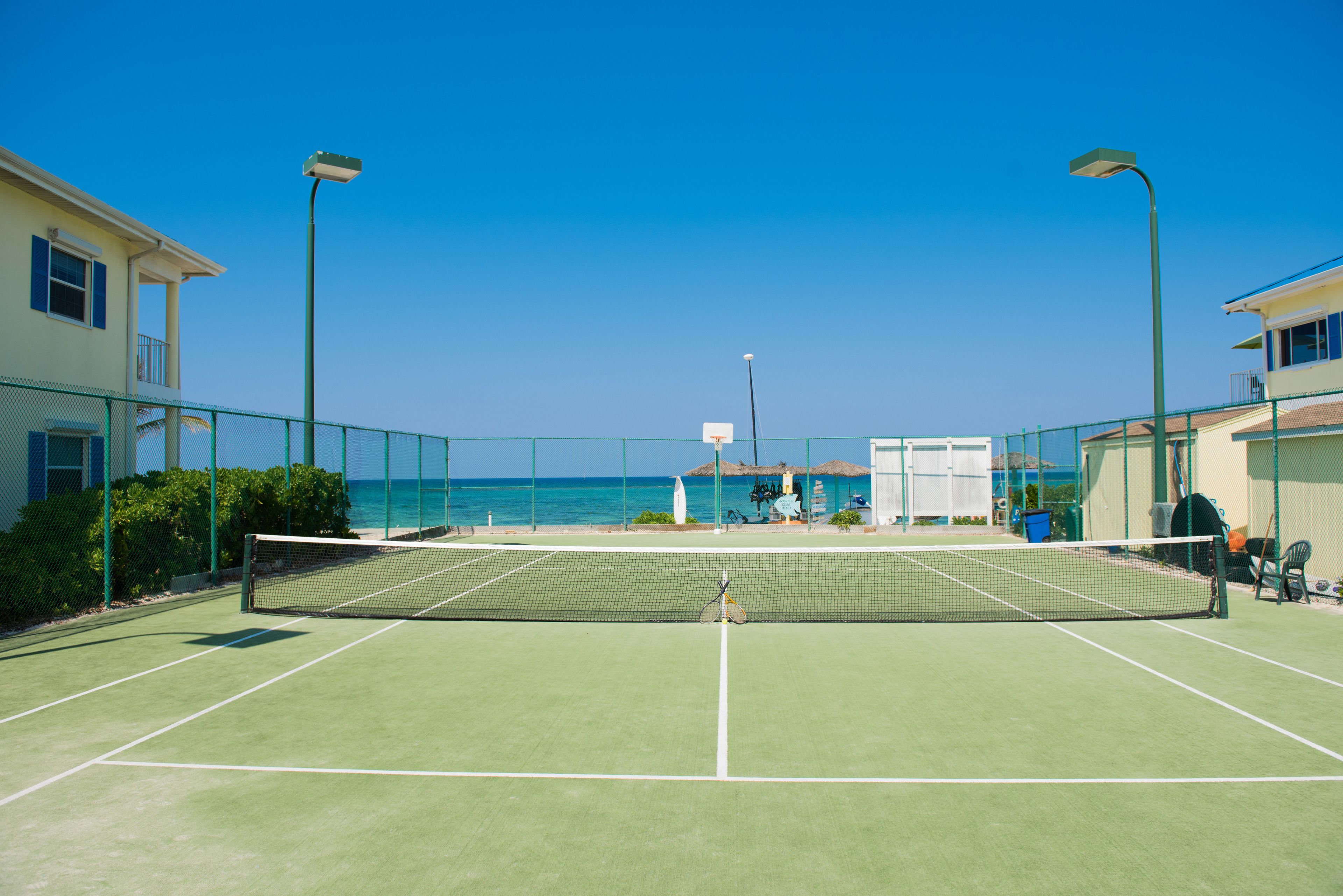 An outdoor tennis court with a net, surrounded by a fence, and overlooking a beach.