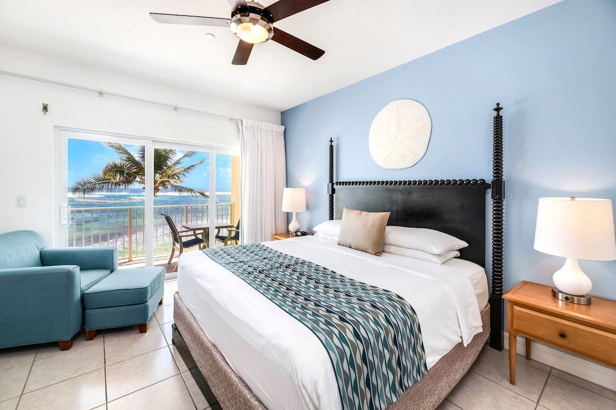 A bedroom with a bed, a ceiling fan, a nightstand, a lamp, a chair, and a view of the ocean through a large window.