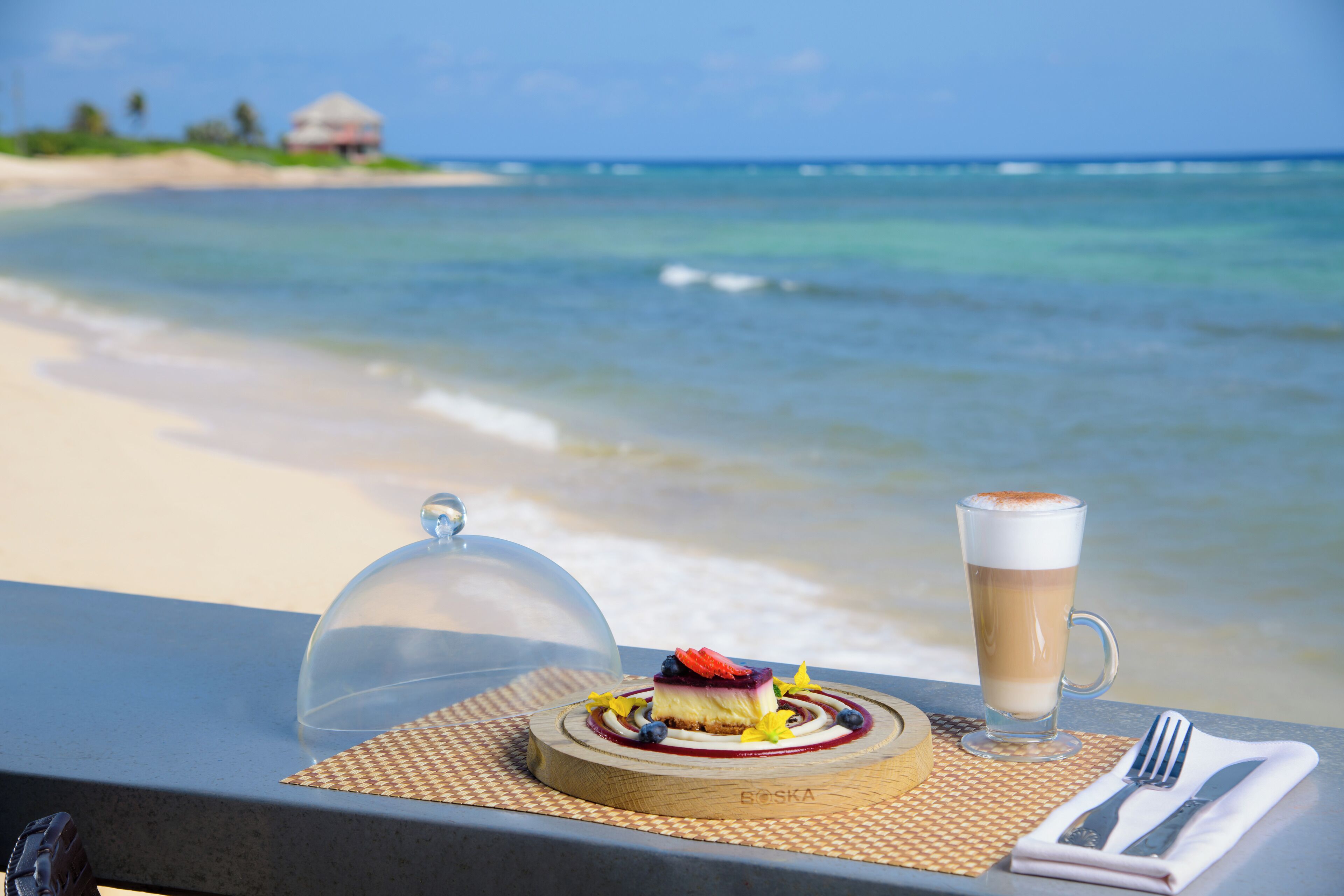 A dessert on a wooden board with a glass of coffee on a table overlooking a beach.