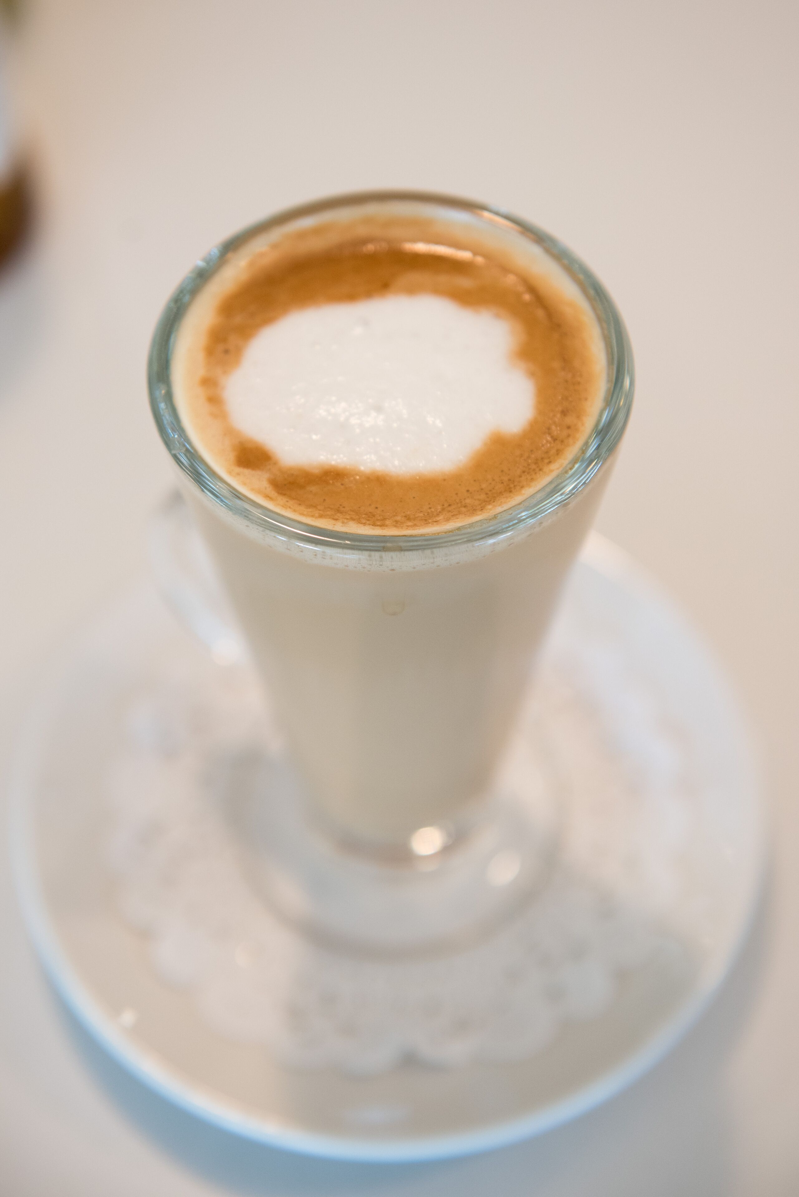 A glass of coffee with a frothy top, placed on a white saucer.