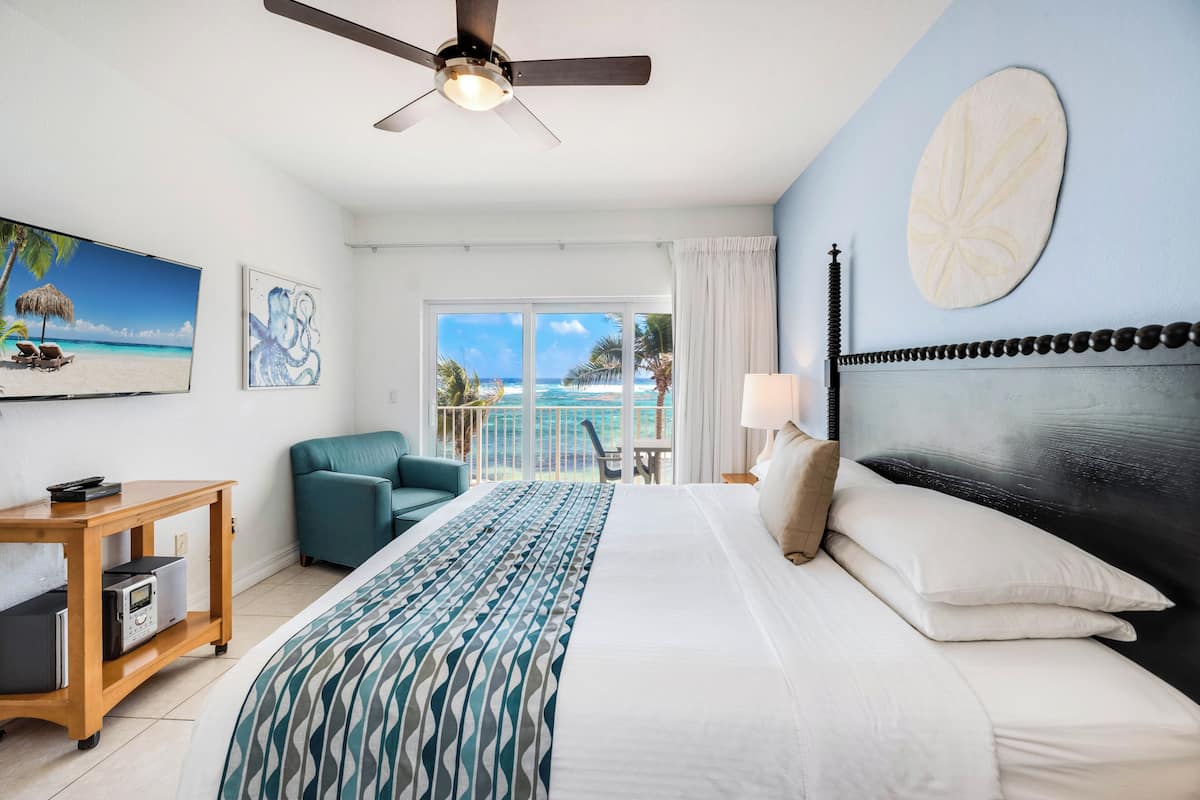 A hotel room with a bed, a ceiling fan, a TV, a view of the ocean, and a balcony.