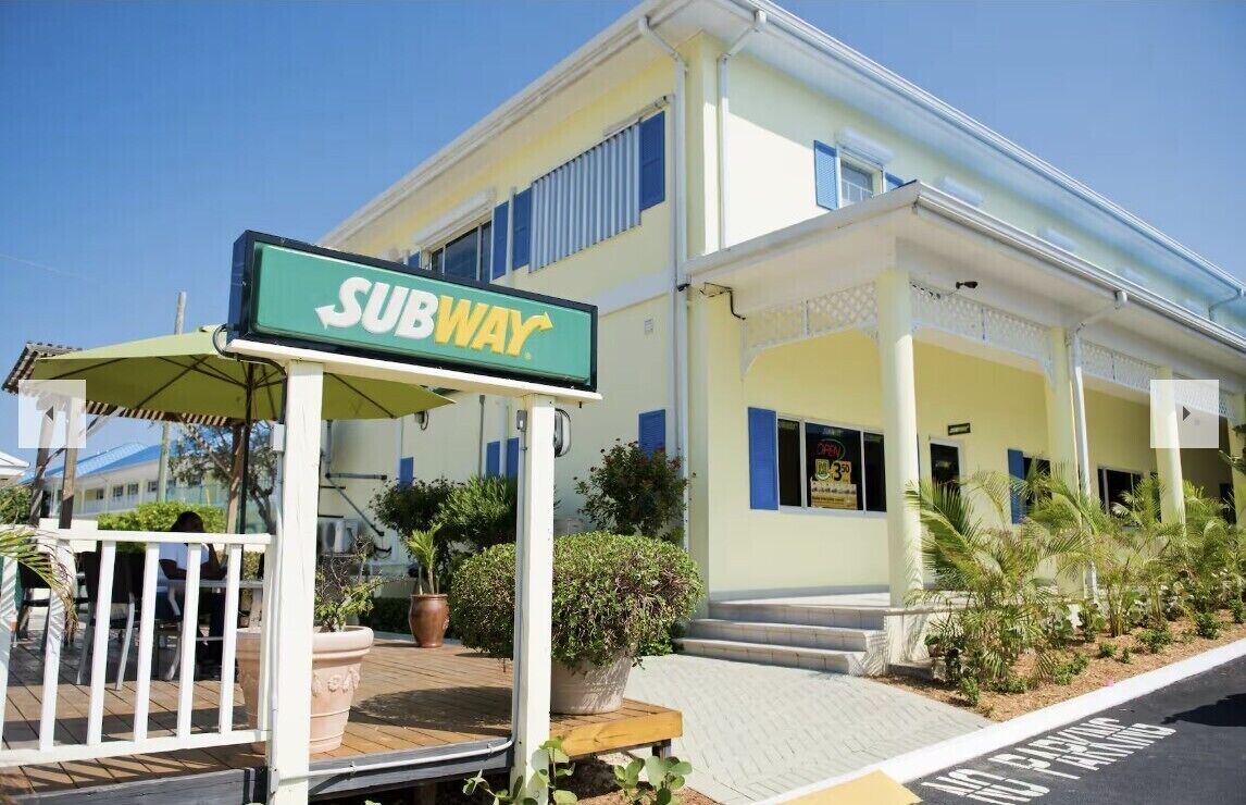 A SUBWAY restaurant with a green sign, outdoor seating, and a building with blue shutters.