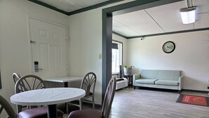 Un comedor con una mesa redonda y cuatro sillas, una puerta blanca, un sofá de color claro y un reloj en la pared.