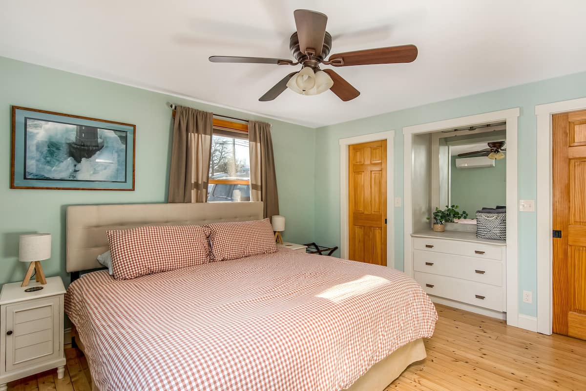 Ein Schlafzimmer mit Bett, Deckenventilator, Nachttisch, Lampe, einem Fenster mit Vorhängen, einer Tür und einem gerahmten Bild an der Wand.