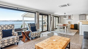 A spacious living room with a large window overlooking a body of water, a wooden coffee table, patterned armchairs, and a dining area with a white table and chairs. - Lakeshore Vermont (Colchester)