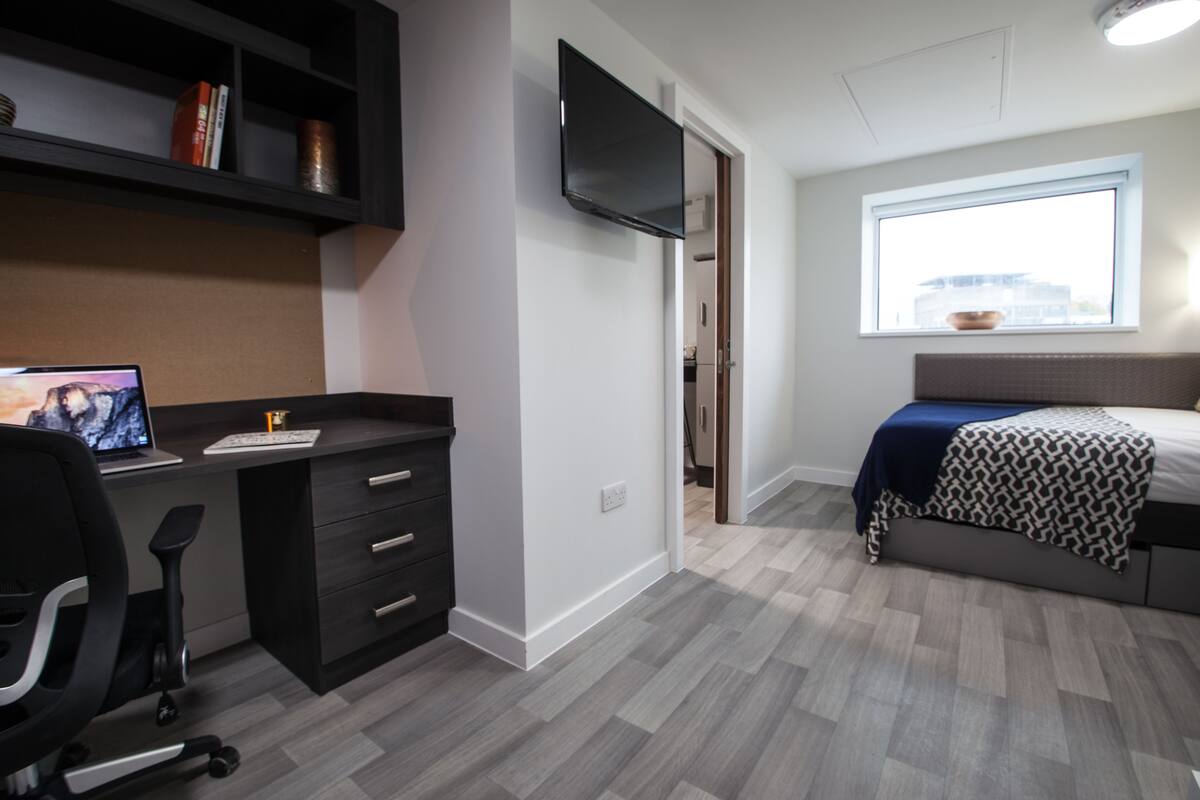 A modern bedroom with a bed, a desk with a computer, a chair, and a TV on the wall.