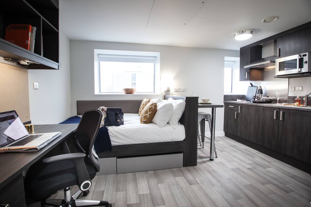 A compact room with a bed, a desk with a laptop, a chair, a kitchenette, and a window.