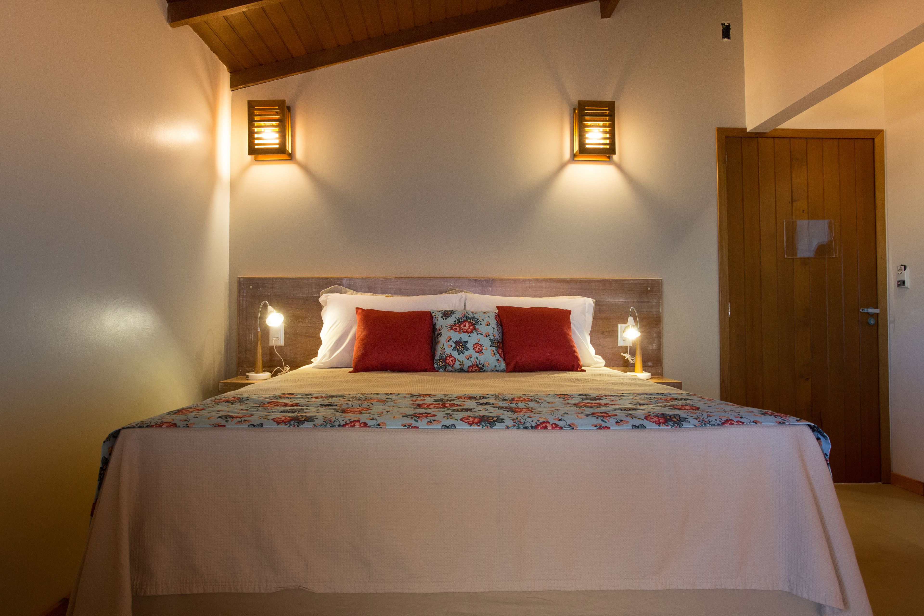 Cama bem arrumada com colcha florida, dois travesseiros vermelhos e duas luminárias na parede.