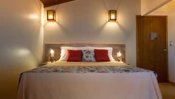 Cama bem arrumada com colcha florida, dois travesseiros vermelhos e duas luminárias na parede.