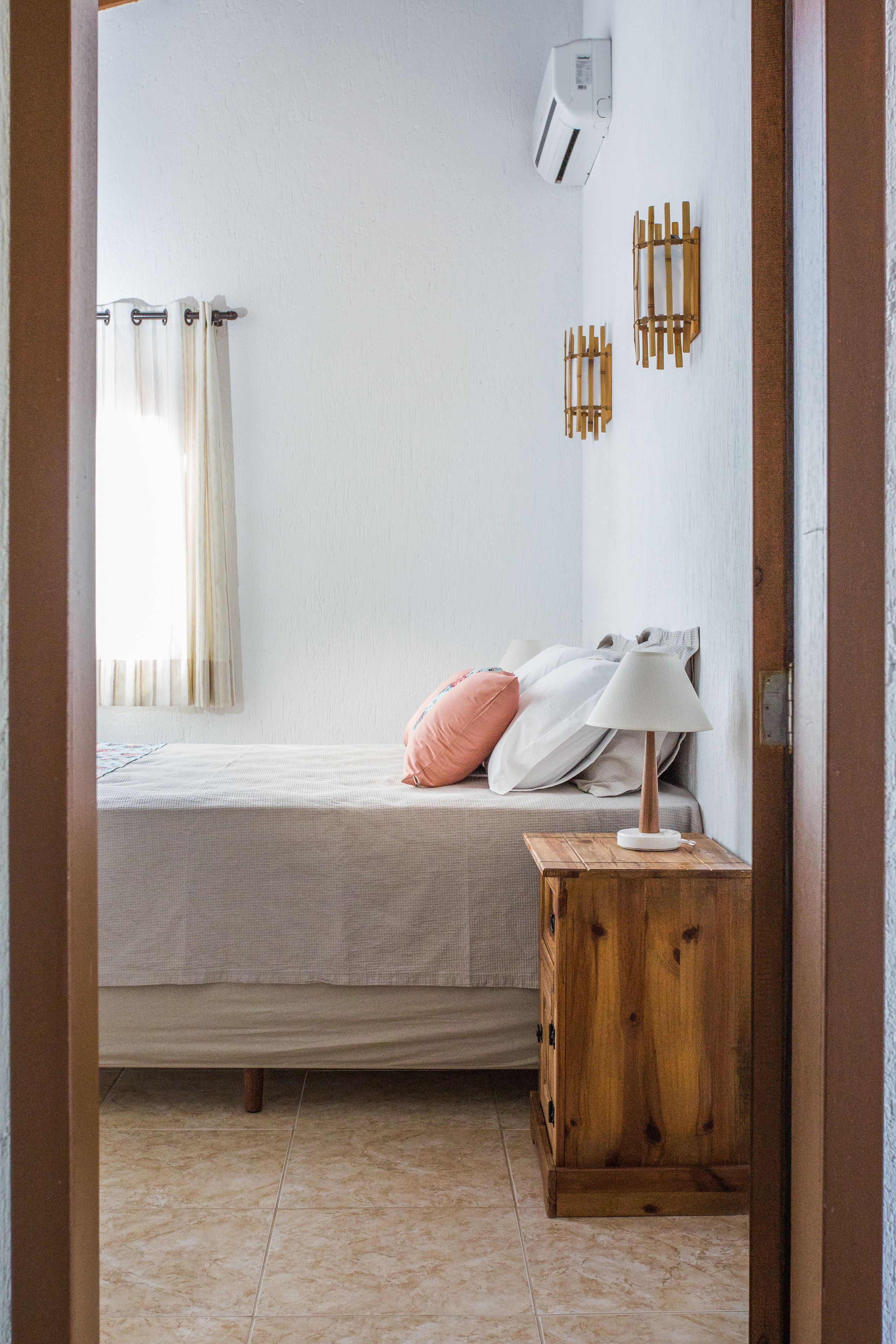 Un dormitorio con cama, mesita de noche de madera, lámpara, calefactor en la pared y ventana con cortinas.