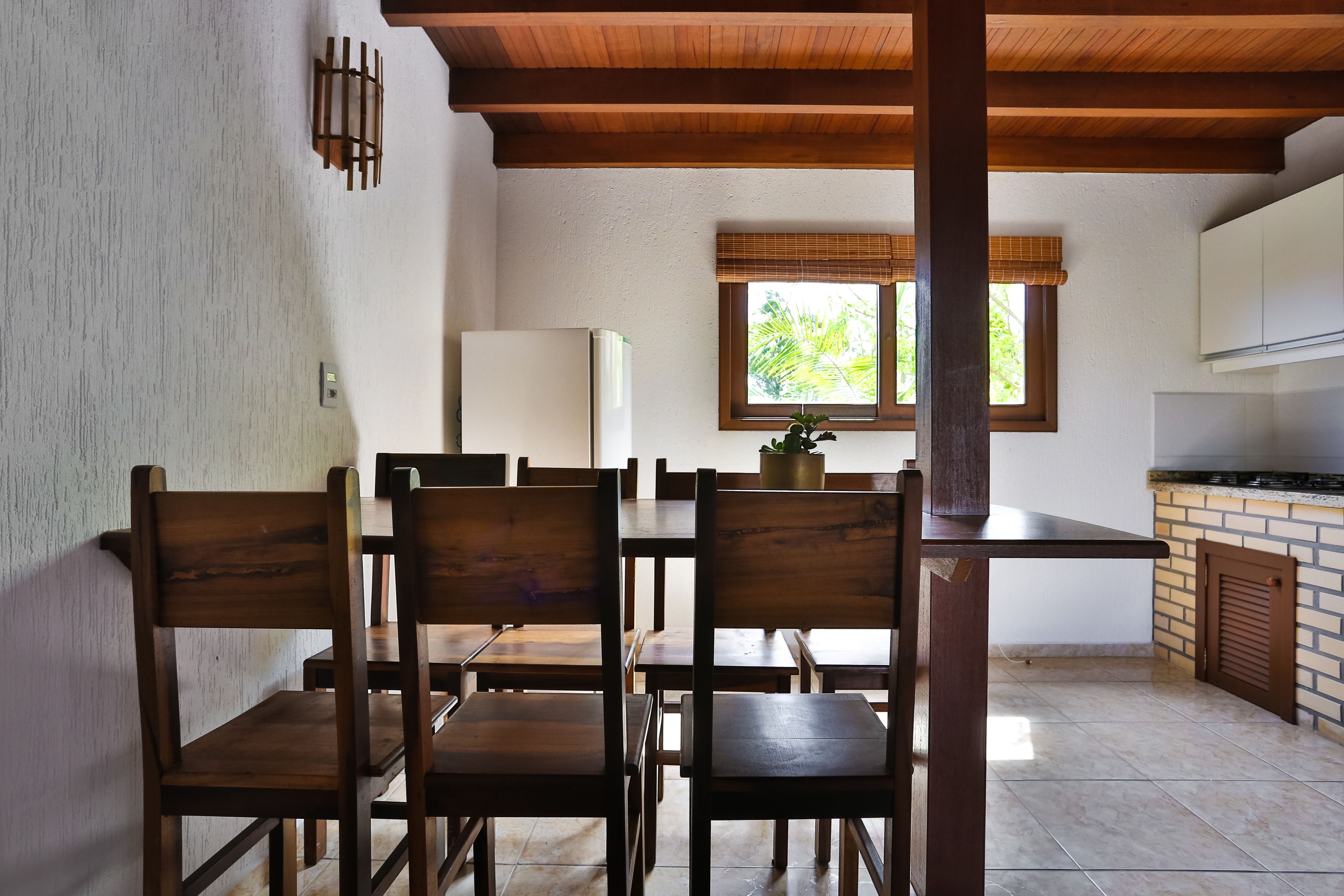 Un coin repas avec des chaises et des tables en bois, un réfrigérateur blanc, une fenêtre donnant sur un espace verdoyant et un coin cuisine avec un mur de fond en briques.