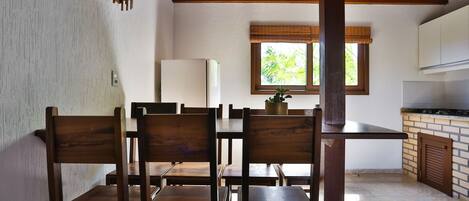 Un comedor con sillas y mesas de madera, un refrigerador blanco, una ventana con vista a follaje, y una zona de cocina con salpicadero de ladrillo.