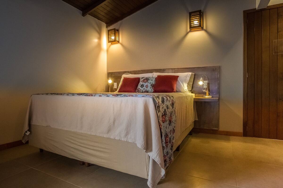 Una cama bien tendida con una colcha estampada con motivos florales y dos almohadas rojas, con lámparas de lectura fijadas en la pared a ambos lados.