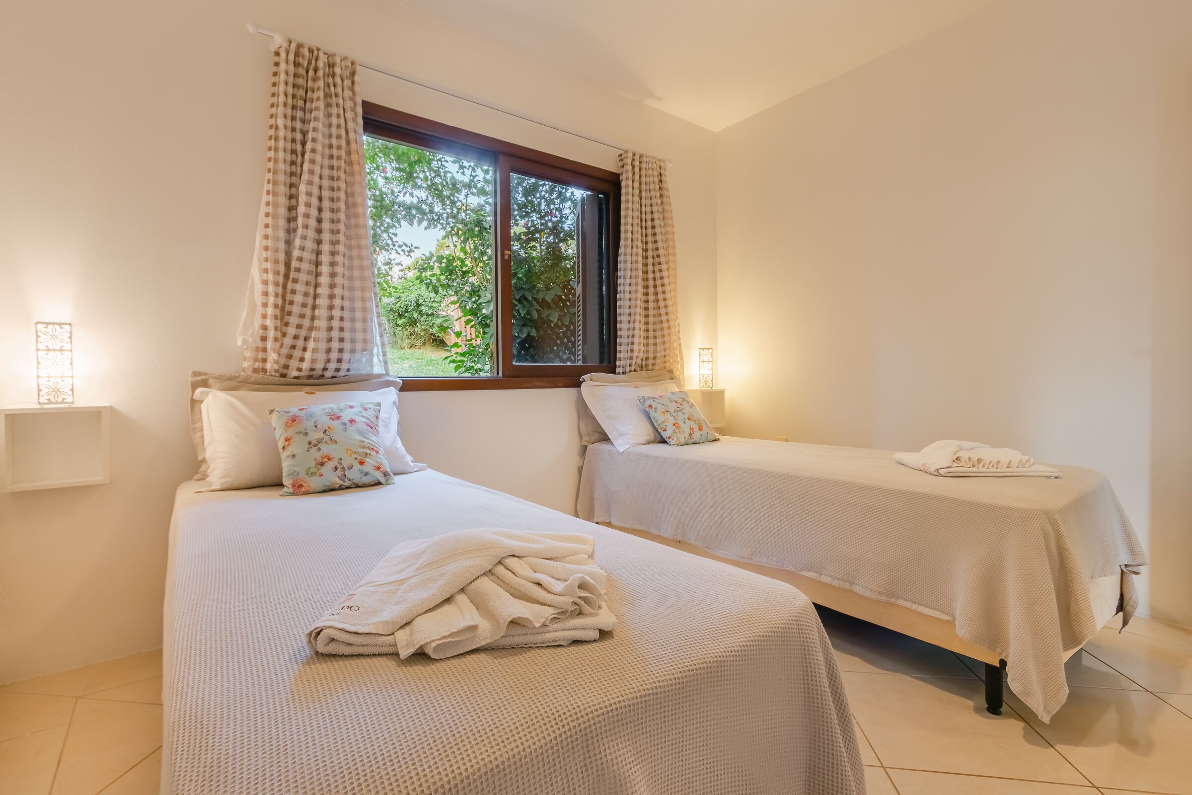 A hotel room with two beds, each with a white bedspread and matching pillows. There is a window with checkered curtains, and a small bedside table with a lamp.