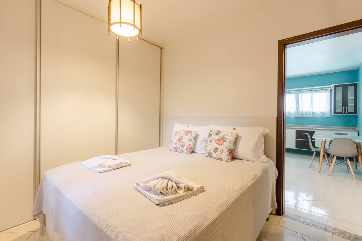 Una cama bien tendida con almohadillas florales y una toalla doblada, una lámpara de noche, y vista a un área de cocina con mesa y sillas.