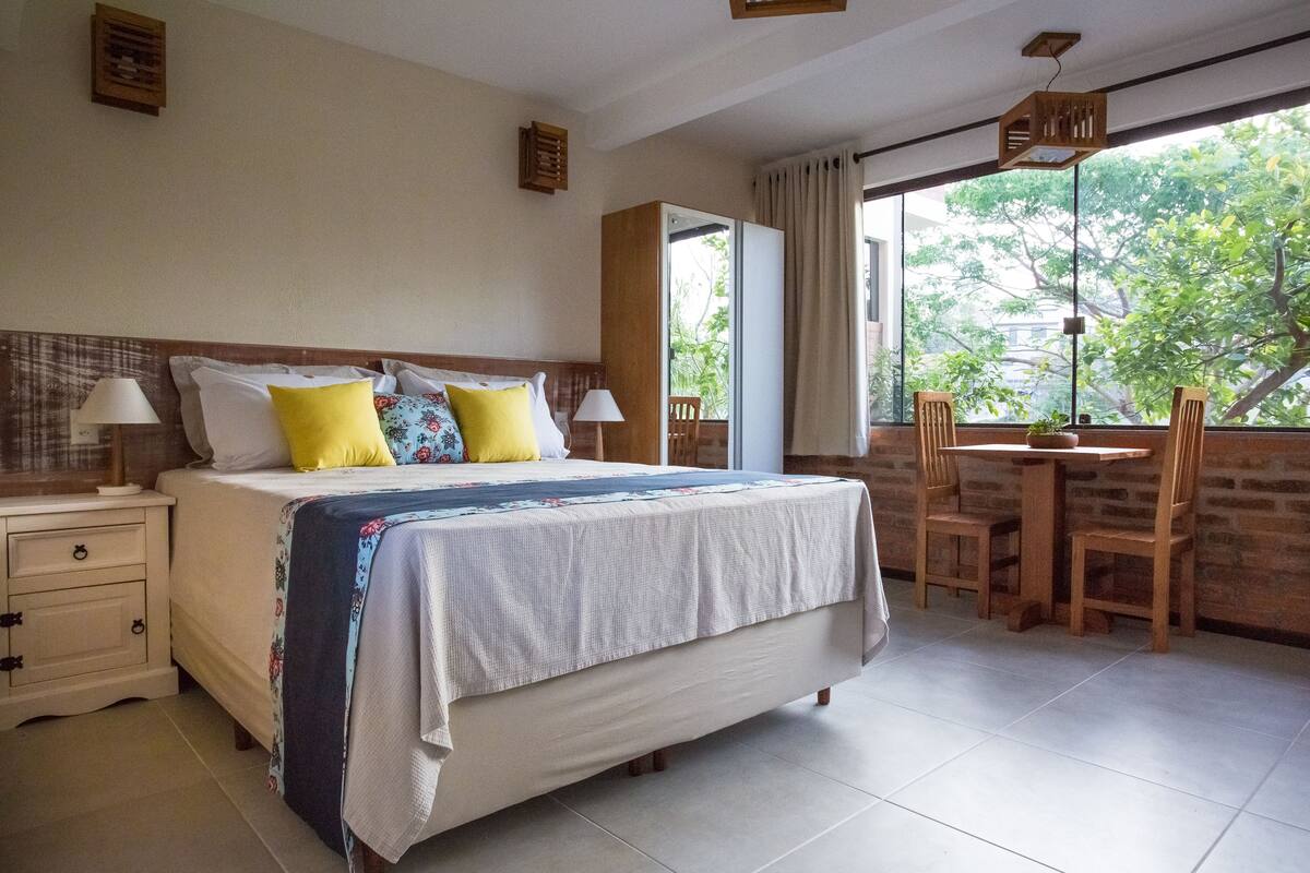 Habitación de hotel con cama, mesitas de noche, una pequeña mesa con sillas y un ventanal con vistas a árboles.