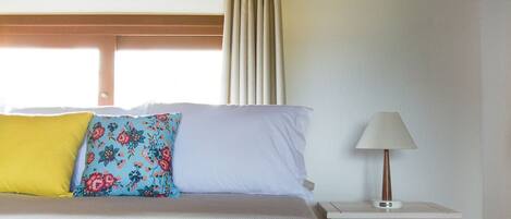 Un dormitorio con techo de madera, una cama con ropa de cama floral, una mesita de noche con lámpara y una ventana con cortinas.
