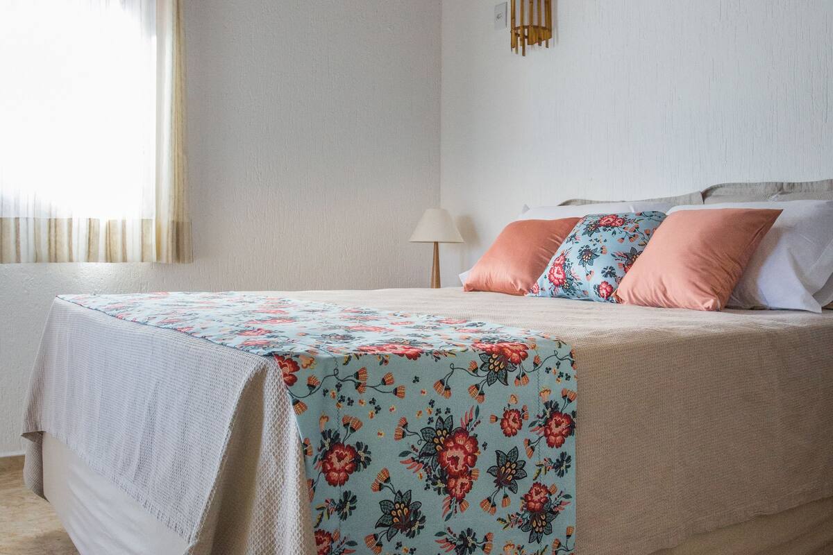 Una cama bien hecha con una colcha estampada, una lámpara de noche y un equipo de aire acondicionado en la pared.