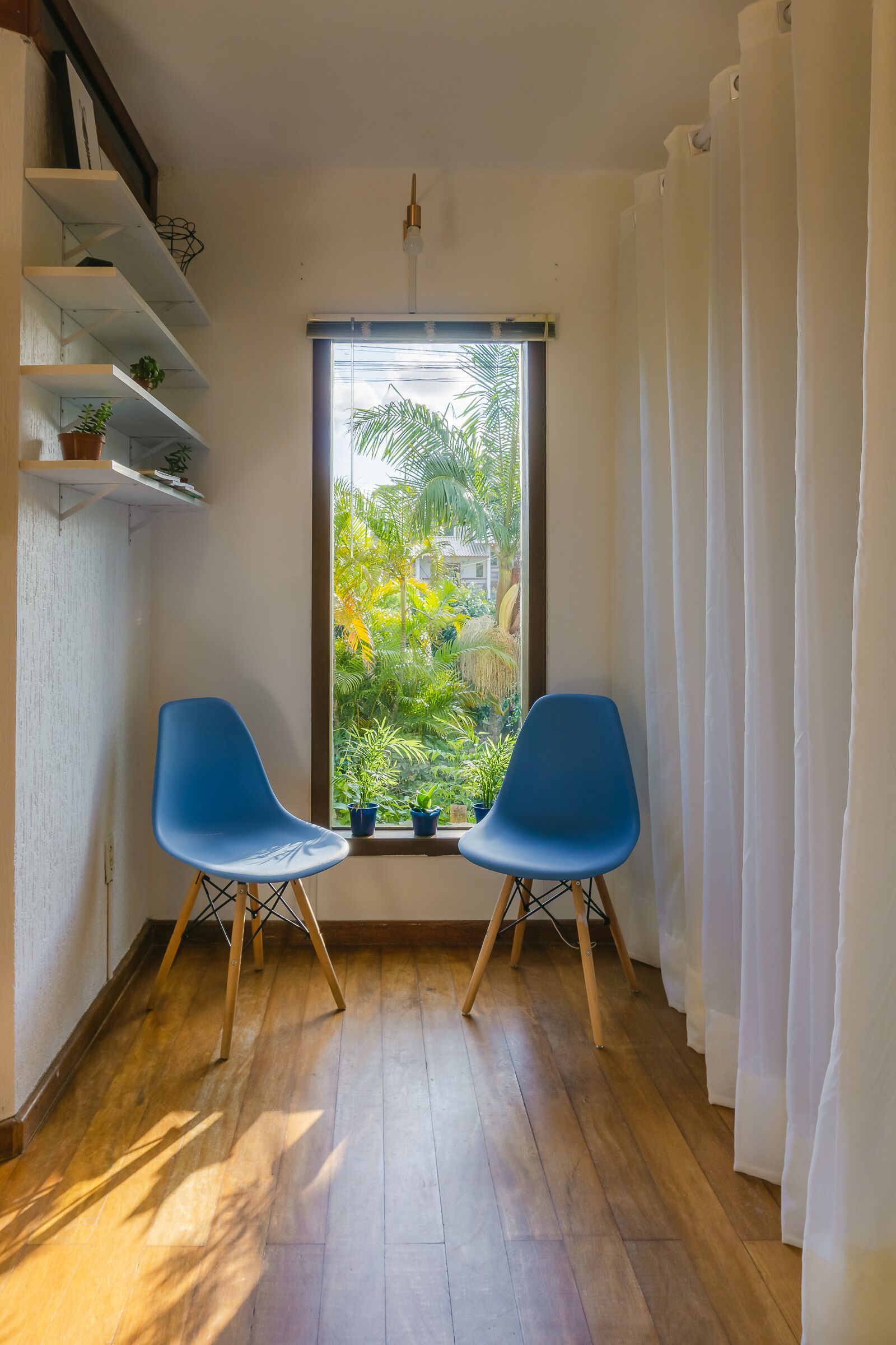 A room with two blue chairs, a window with a view of greenery, and a white curtain.