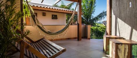 A hammock chair and wooden tables in a covered outdoor area with a view of a house and greenery.
