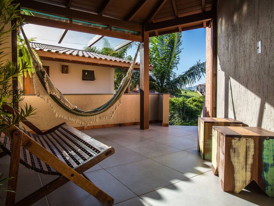 A hammock chair and wooden tables in a covered outdoor area with a view of a house and greenery.