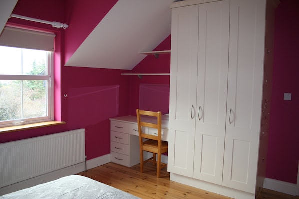 A bedroom with pink walls, a wooden floor, a white radiator, a bed, a desk, and a wardrobe.