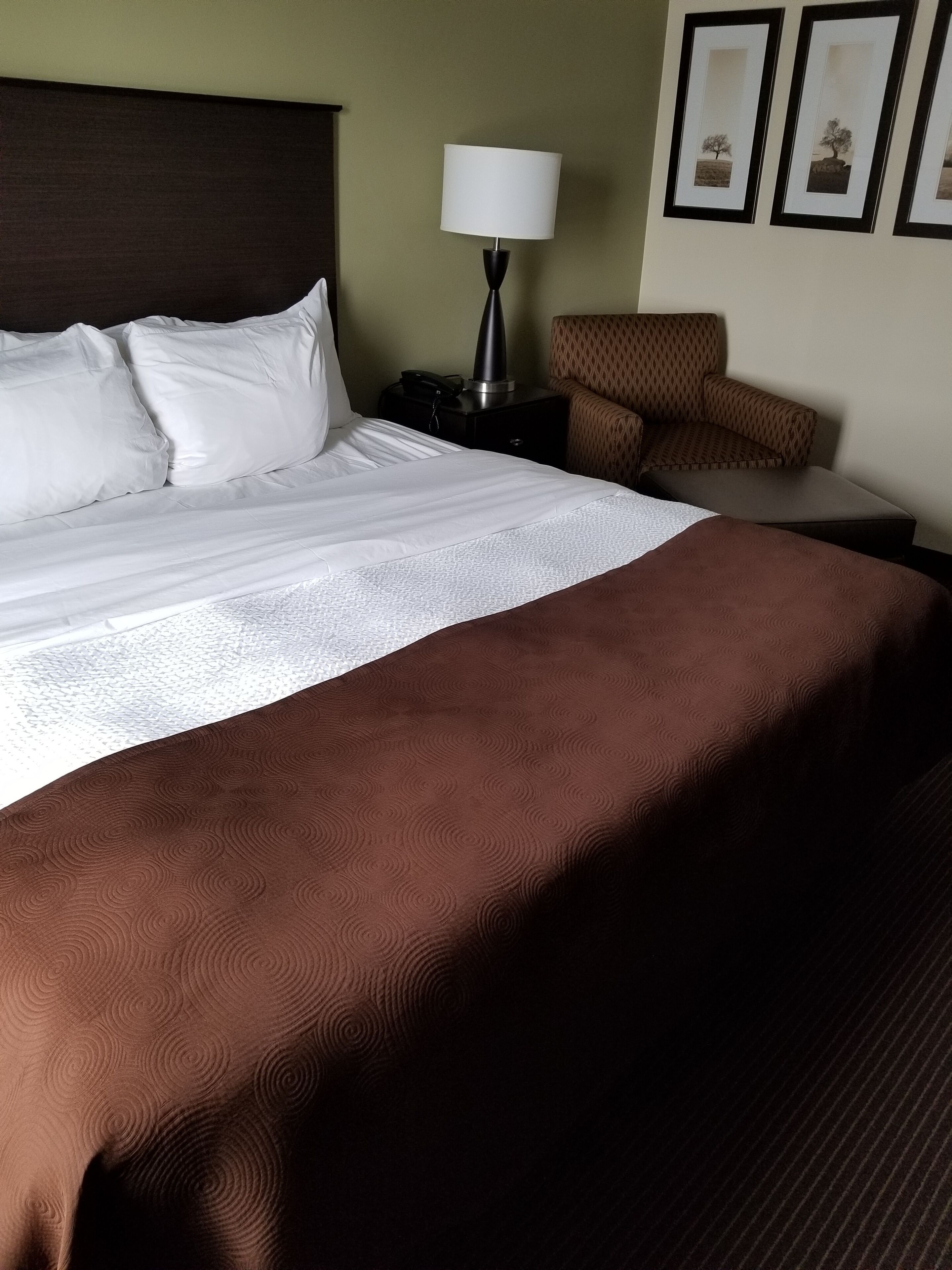 A neatly made bed with white and brown bedding, a bedside lamp, a patterned armchair, and framed pictures on the wall.