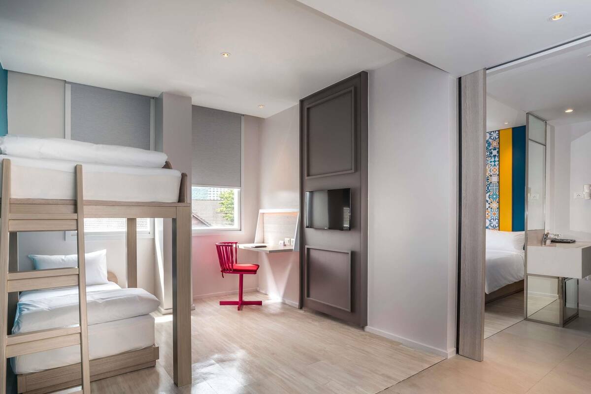 Una habitación de hotel moderna con una cama litera, un escritorio con una silla, un televisor y un baño visible a través de una puerta abierta.
