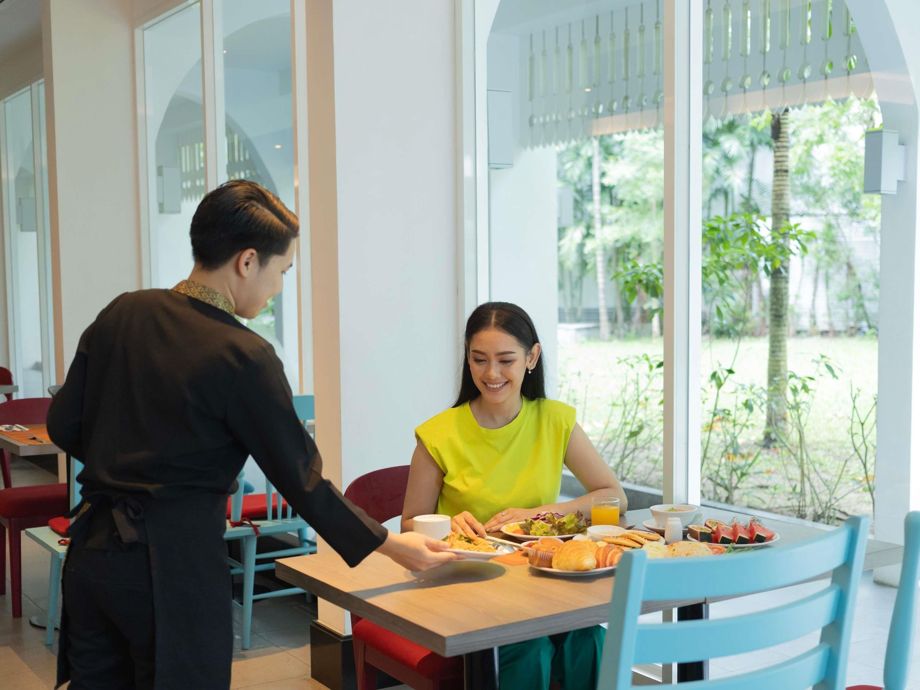 A dining area with a table set for two, a waiter serving food, and a view of greenery outside.