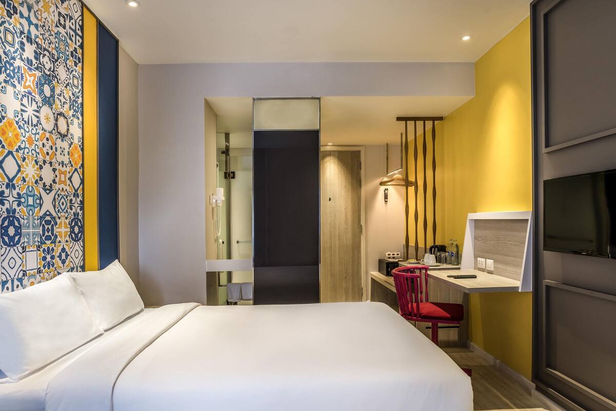 Habitación de hotel con cama, escritorio con silla, televisor y baño visible a través de una puerta abierta.