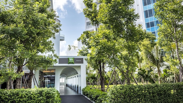 Une entrée d’hôtel moderne, dotée d’une galerie couverte, entourée d’une végétation luxuriante et de grands immeubles aux fenêtres teintées de bleu.