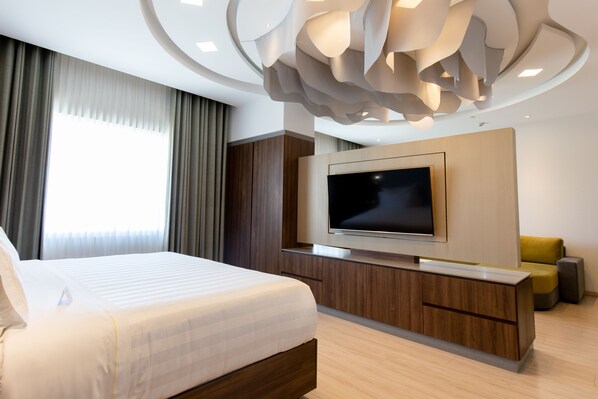 Una habitación de hotel moderna con una cama grande, un televisor de pantalla plana montado en una consola de madera, un sofá y una lámpara de techo original.
