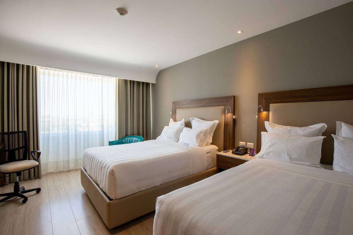 Ein Hotelzimmer mit zwei Betten, einem Schreibtisch mit Telefon, einem Stuhl und einem großen Fenster mit Vorhängen.