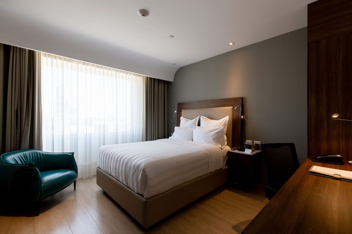 Ein modernes Hotelzimmer mit einem großen Bett, einem Schreibtisch, einem Sessel und Blick auf die Stadt durch transparente Vorhänge.